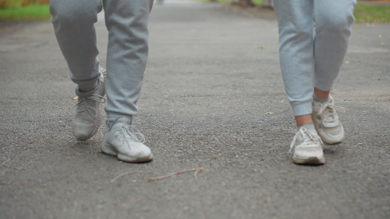 Leg view of individuals wearing grey joggers and sneakers walking side by side on tarred road with blurred background of greenery and distant figure