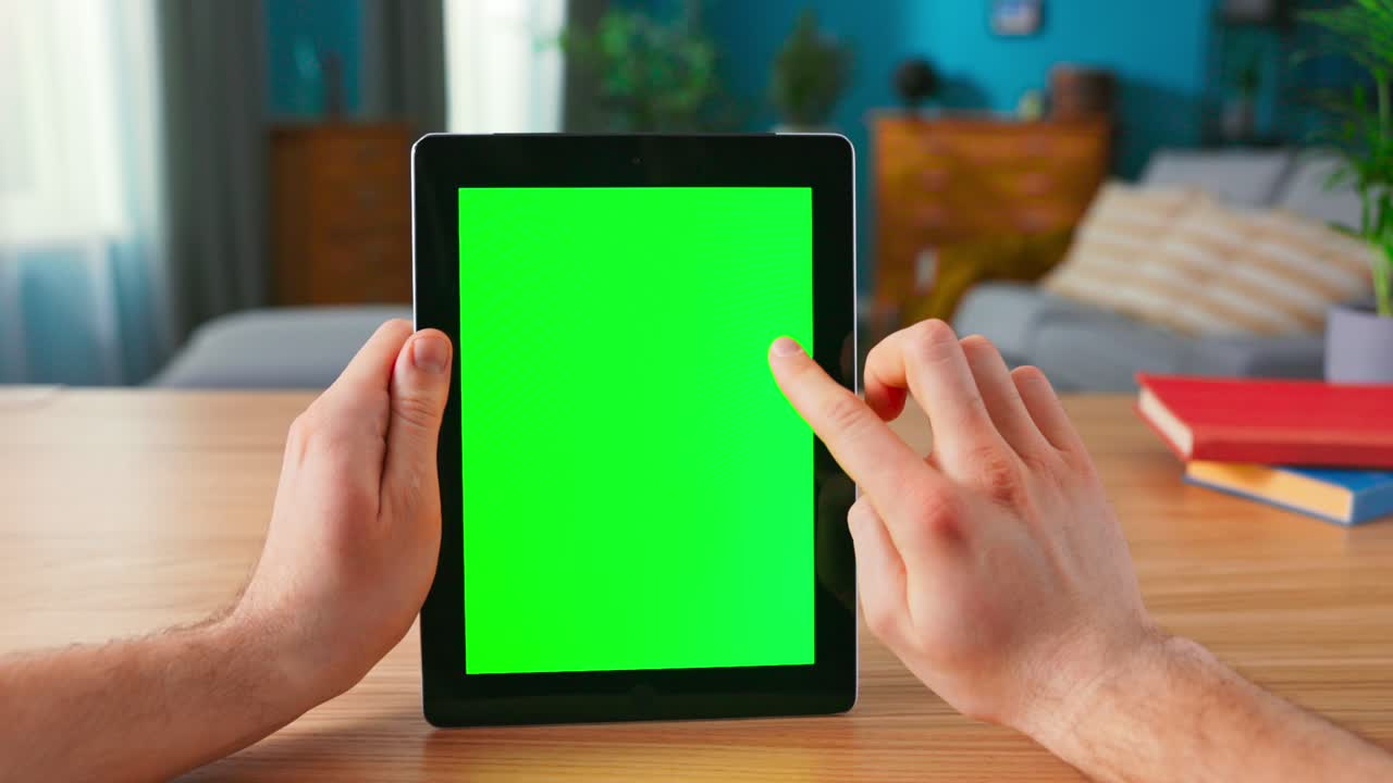 primer plano de un hombre usando gestos con las manos en una pantalla de maqueta verde, una tableta digital en modo paisaje mientras está sentado en su escritorio. en el fondo, una acogedora sala de estar