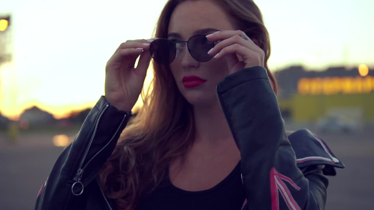 joven mujer encantadora con labios rojos y cabello largo en chaqueta de cuero lleva gafas de sol