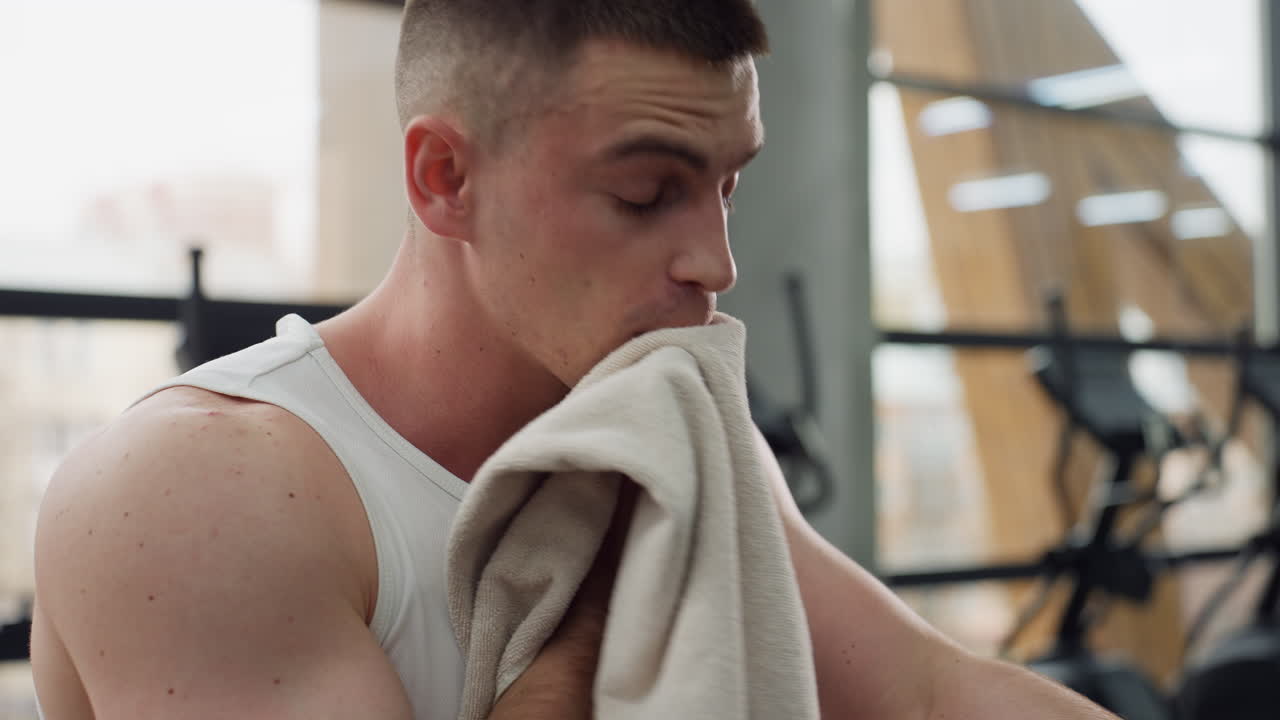 Close up of sports man cleaning sweat off body and face with soft towel after intense elliptical workout, inhaling deeply in satisfaction as muscles cool down in bright modern indoor gym