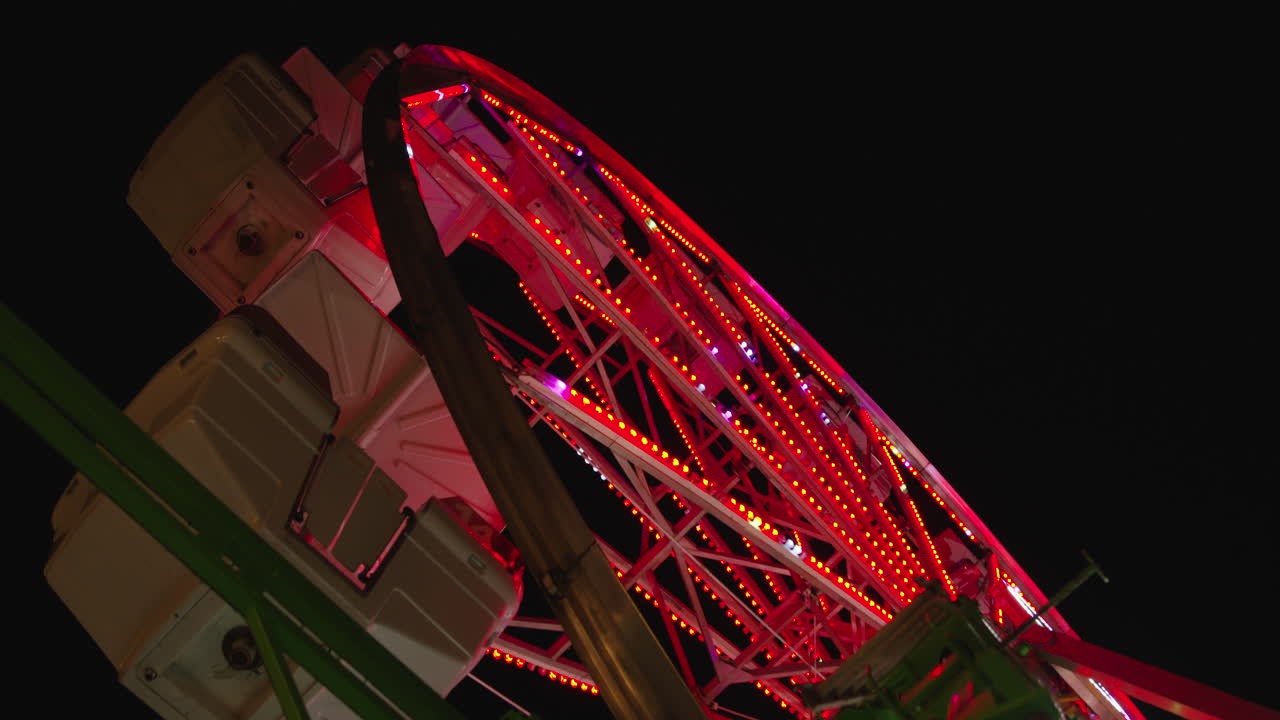 una toma en ángulo bajo de una rueda de la fortuna se inclina hacia arriba para revelar la parte superior mientras las góndolas se balancean con el viento por la noche y las luces de la rueda de la fortuna cambian de color