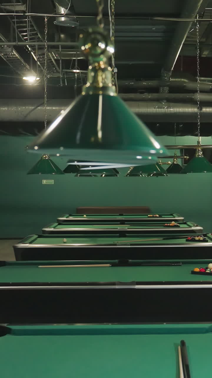 Immagine di tavoli da biliardo verdi con le bilie disposte e stecche appoggiate. Lampade a sospensione forniscono un'illuminazione soffusa, creando un'atmosfera accogliente nella sala da biliardo.