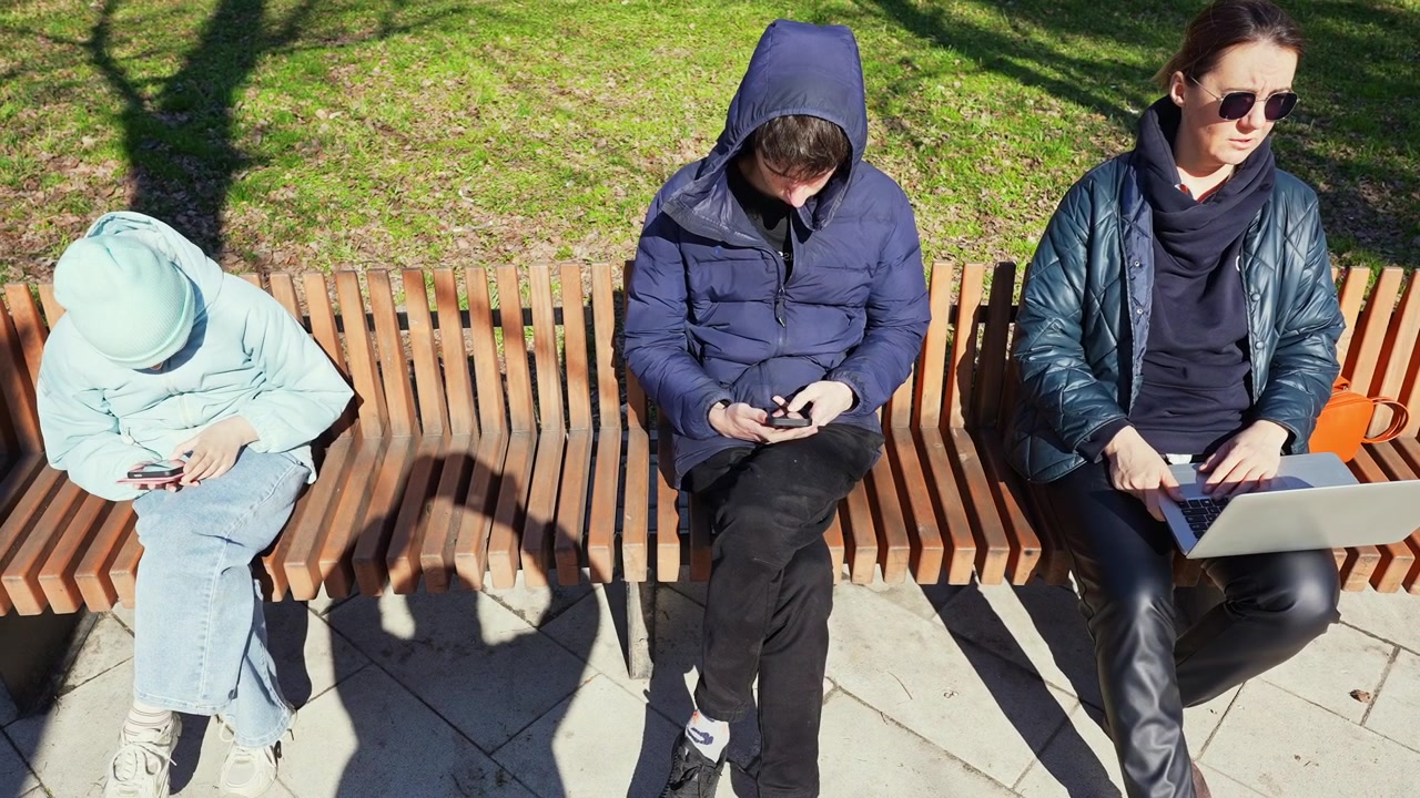 Kolme ihmistä käyttää digitaalisia laitteita puistonpenkillä istuen