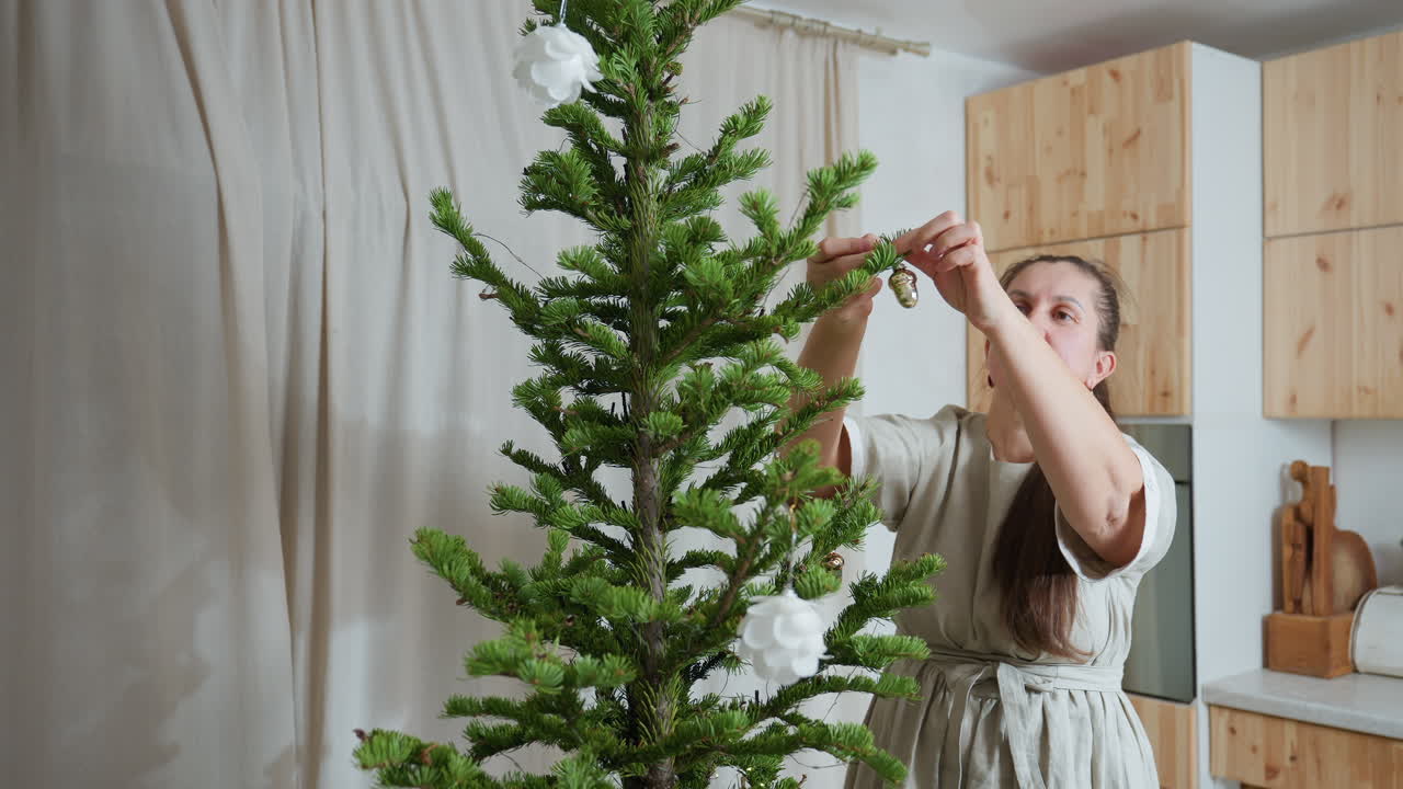 Woman in casual gown fixing ornaments on christmas tree with white flower decorations and string lights in cozy kitchen with natural wooden cabinets, preparing festive atmosphere