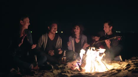 multiethnic group of young people sitting by the bonfire late at night and singing songs and playing guitar, clapping hands