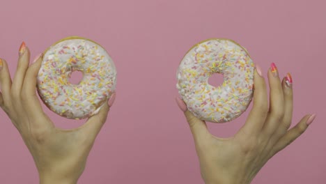 las manos de las mujeres sosteniendo dos rosquillas blancas, deliciosas y salpicadas sobre un fondo rosa.