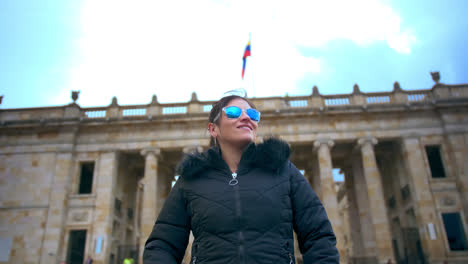 medium shot of a woman dressed with black jacket and blue sunglasses stare at the government palace in bogotá in a sunny afternoon-1