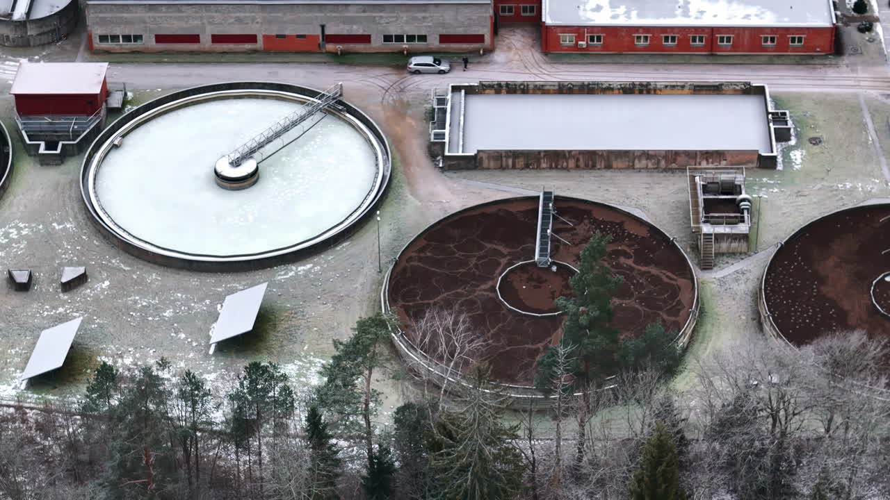 Aerial of wastewater processing circular industrial tanks surrounded by snowy landscape and trees, static overview