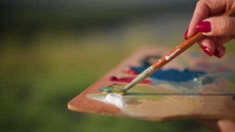 a close-up shot of a hand holding a wooden palette while using a small brush to mix vibrant colors. the scene is set outdoors in natural sunlight