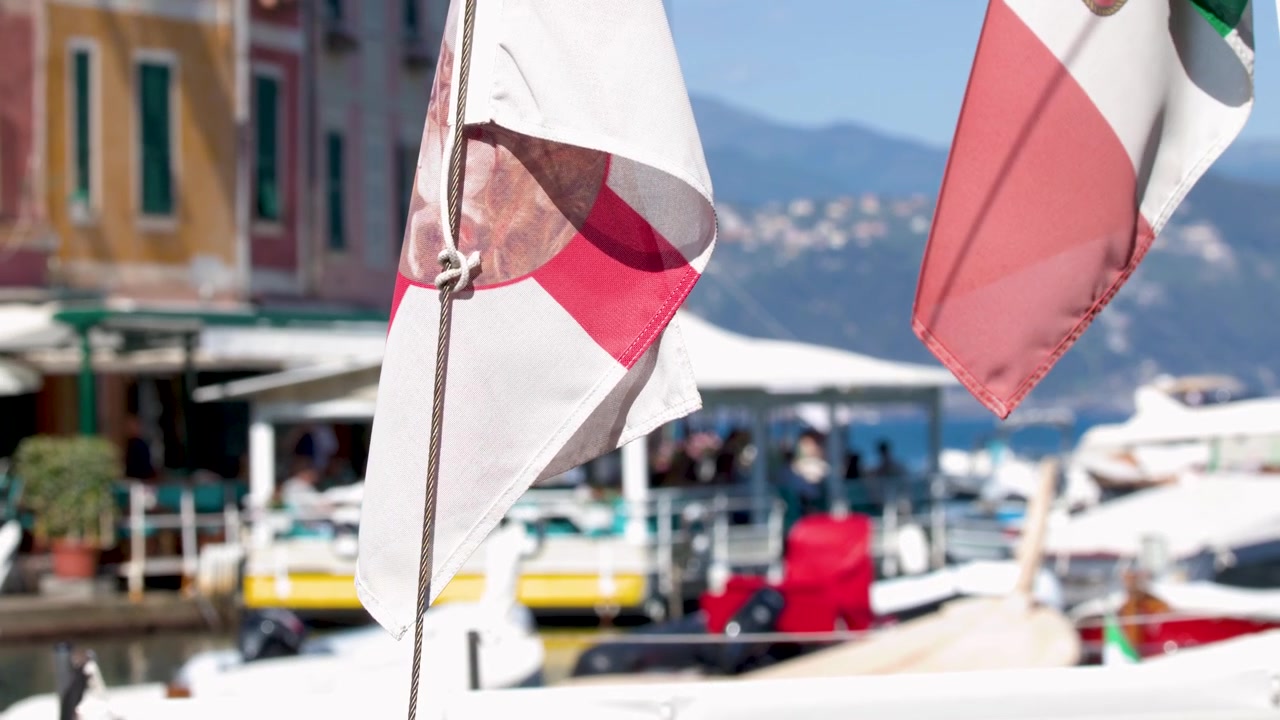 Primo piano di una bandiera italiana che sventola su un soleggiato porto turistico mediterraneo con architettura vivace.