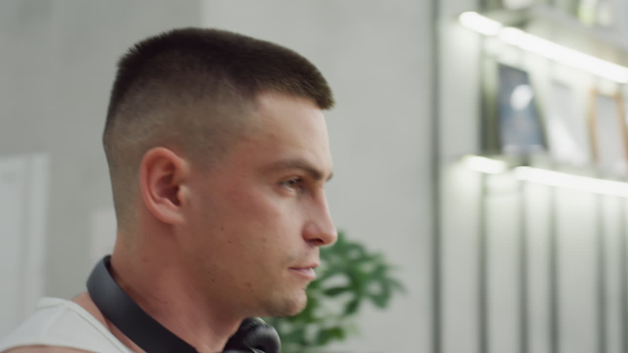 Fitness enthusiast wearing headphones around neck walks toward gym equipment station in modern fitness center with blurred exercisers and industrial interior under bright natural light