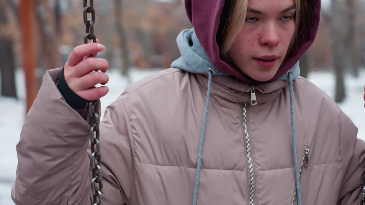 крупный план молодого взрослого человека, хватающего цепь для качелей во время подъема для баланса, в теплом зимнем пиджаке с капюшоном, покрытой снегом земле и размытыми фигурами на заднем плане