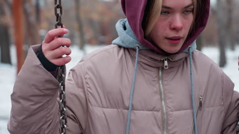 close-up of young adult gripping swing chain while climbing for balance, wearing warm winter jacket with hoodie, snow-covered ground and blurred figures in background