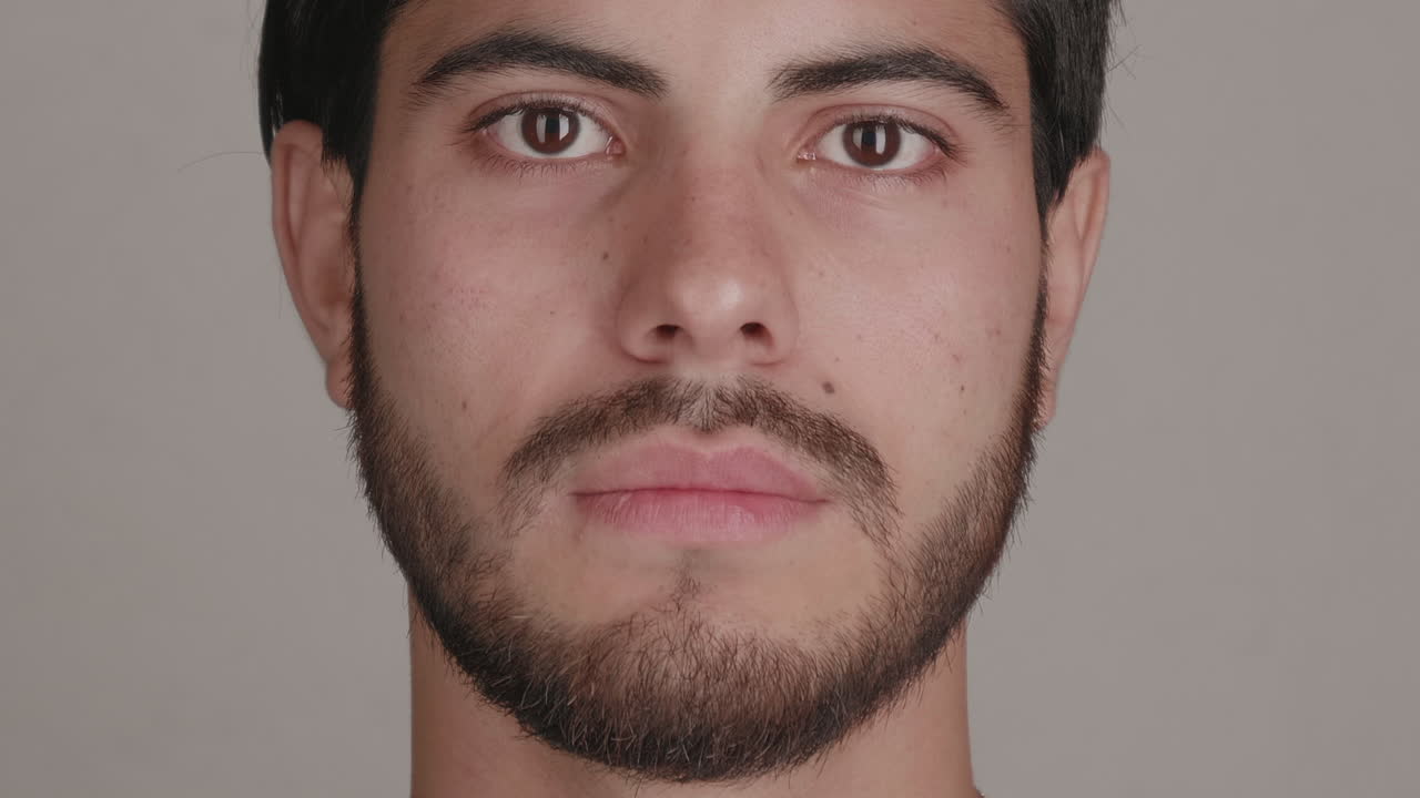 High-quality 4K footage of a diverse young man with facial hair looking straight into the lens against a grey background. Ideal for modern casting, men's health, and identity concepts