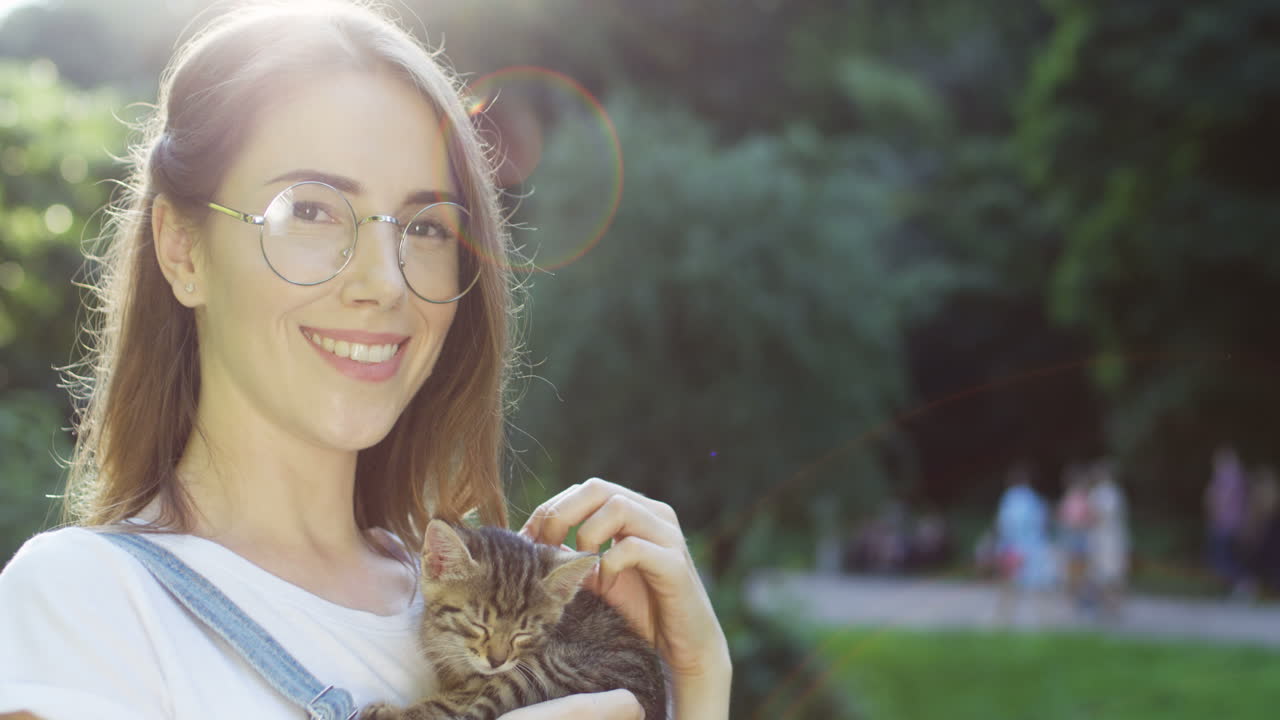 Close-up view of a caucasian woman in glasses holding and petting kitty cat while smiling at camera in the park on a summer day