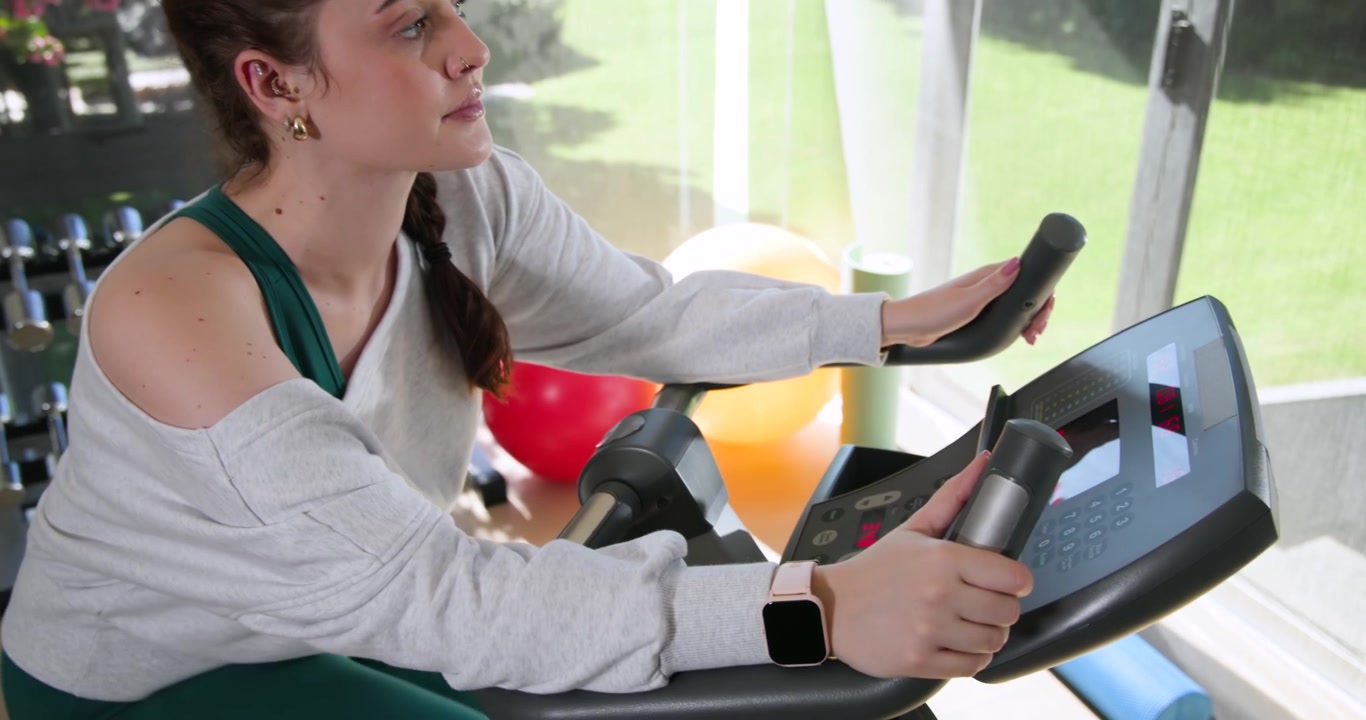 Mujer con top de gimnasio sentada en una máquina de cardio, presionando la consola para comenzar el entrenamiento y mirando por la ventana.