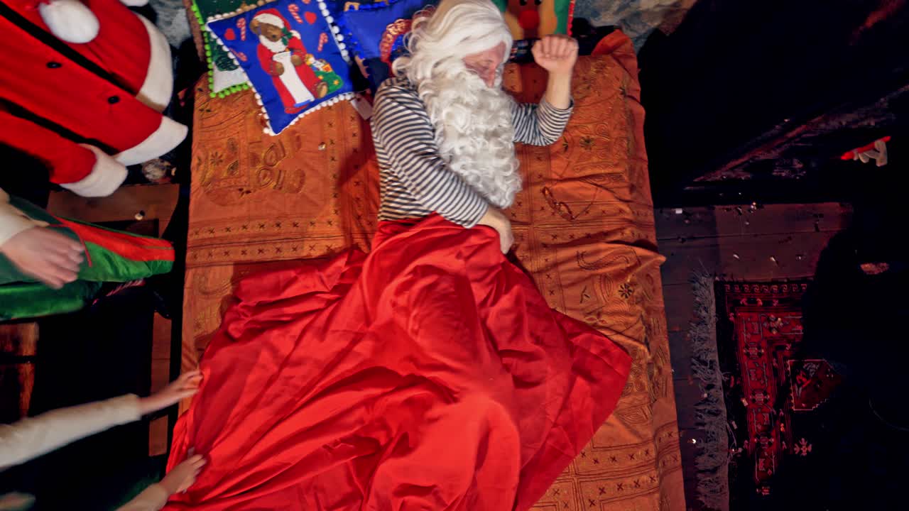 Santa Claus is sleeping in specially decorated room in his bed. Elves are waking him with a ringing a bell and by touching him. Christmas and New year concept close up video. Video from above.