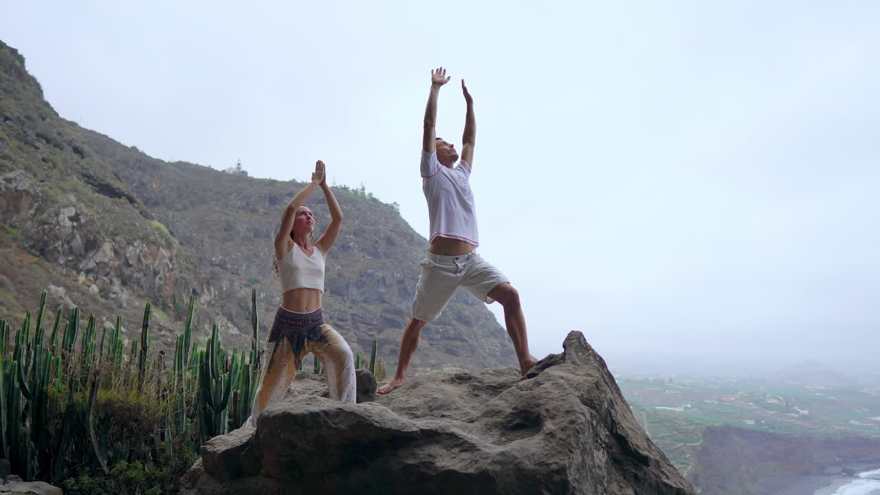 With the ocean below, a man and woman on a cliff's edge raise their hands and inhale the sea air during yoga, absorbing its serenity