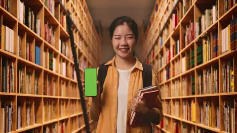 asian woman student with a backpack and some books smiling and showing green screen smartphone to camera while standing in the library