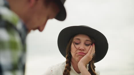 a close-up shot of a woman wearing a hat and white dress, displaying an exhausted expression with her hand on her face. in the blurred background, a man is partially visible