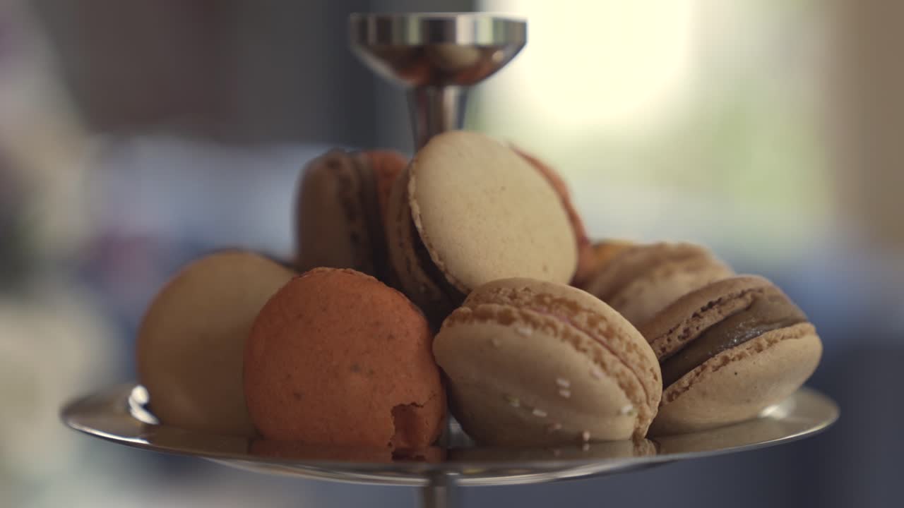 Close-up of assorted beige and brown macarons displayed on a silver stand in soft natural light, with a blurred indoor background creating an elegant and inviting atmosphere.