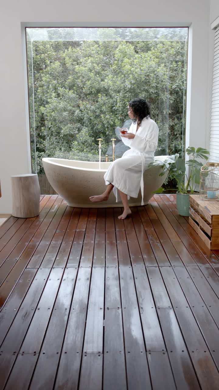 Vertical video of biracial woman sitting on bathtub drinking tea at home, slow motion