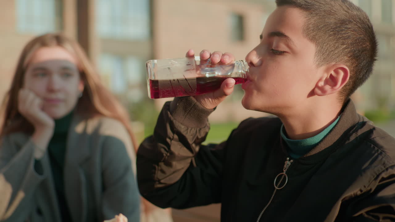 Kid eating sausage while holding juice and sipping drink as woman in background looks at him with admiration, both sitting outdoors at wooden table under warm sunlight