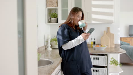 happy plus size biracial female doctor with coffee using smartphone at home, copy space, slow motion