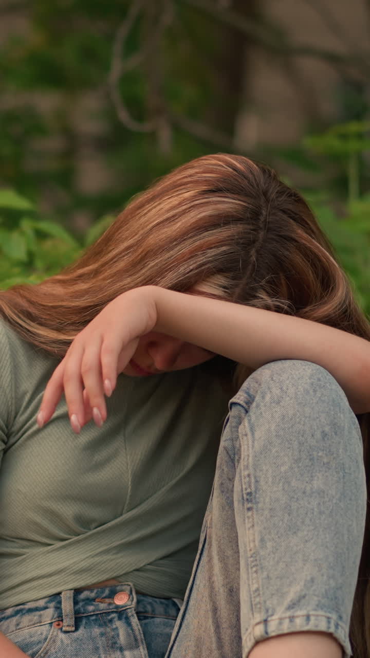 Woman in green top and jeans sits on wooden bench in park, resting her head on her arm in a reflective, somber posture, surrounded by lush greenery and trees, she conveys a peaceful solitude