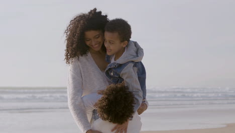 joven madre estadounidense sosteniendo a su hijo y acariciando el cabello de su hija mientras pasa tiempo en la playa en un día soleado