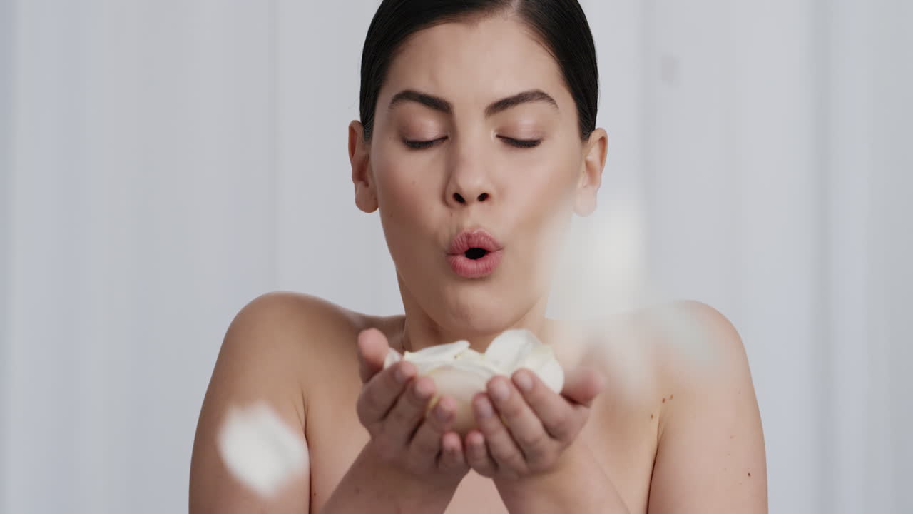 천천히 움직이는 초상화 아름다운 젊은 백인 여성이 장미 잎을 불어 고급스러운 피부 관리 향수를 즐기고  ⁇ 색 배경에 부드러운 건강한 피부를 가진 매력적인 여성