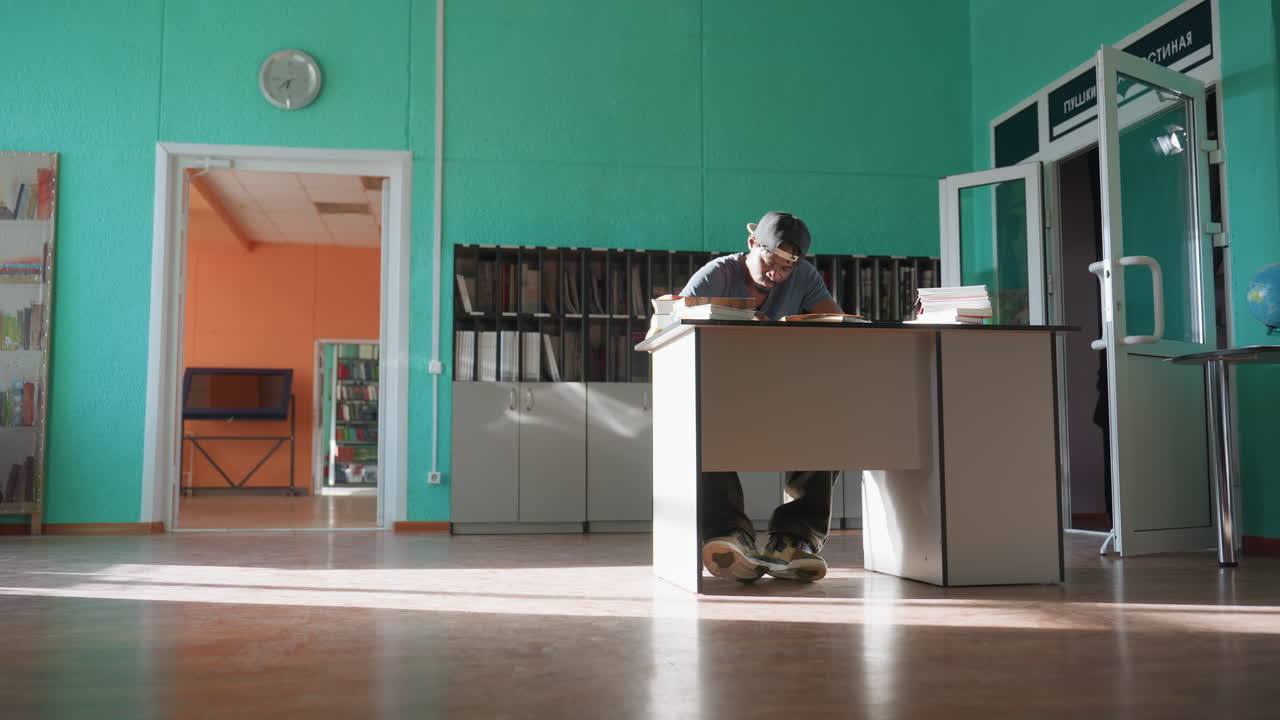 Student sits at desk in brightly lit library, focused on writing in notebook with pen, surrounded by stacks of books as sunlight streams across polished floor