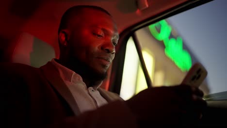a confident man with black skin and a brown suit works using his white smartphone and looks around while sitting in the passenger seat in the interior of a modern car. the bright red light of the evening city falls on the face and interior of the car