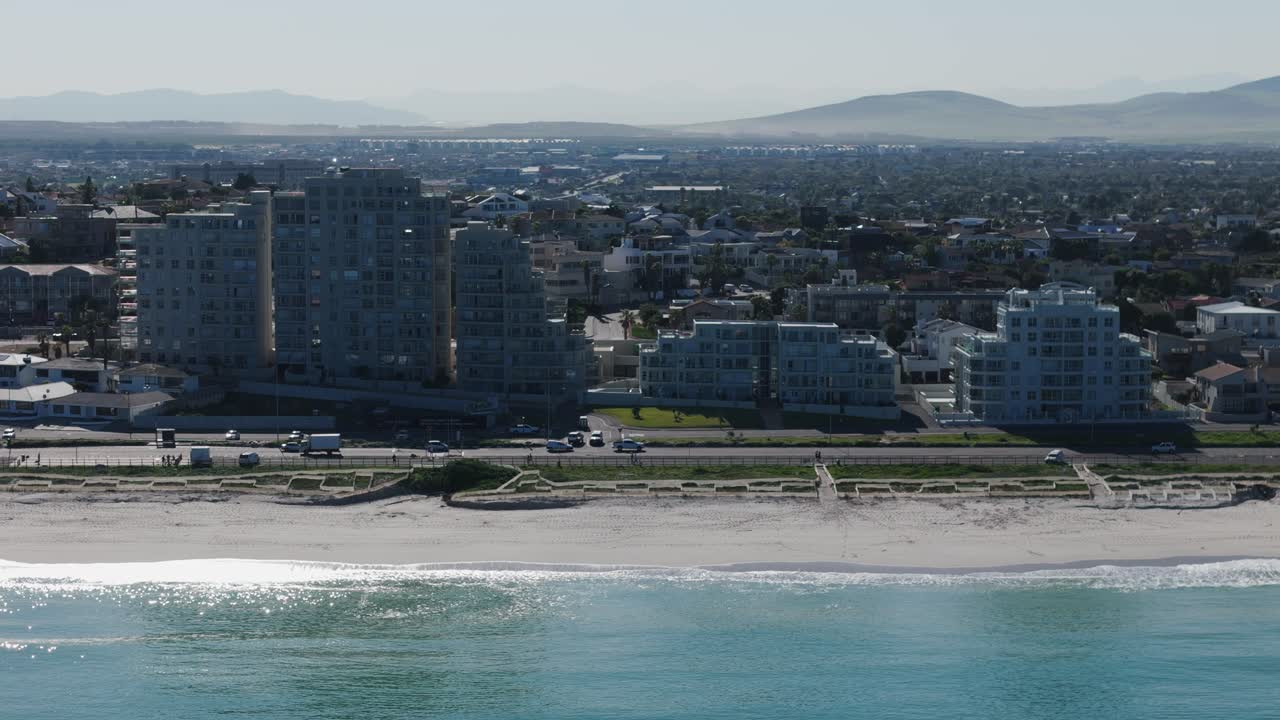 바가 마을의 드론 클립으로 바쁜 해변 도로와 조용한 해변을 보여 주고, 배경에는 아파트 건물과 집이 있습니다.