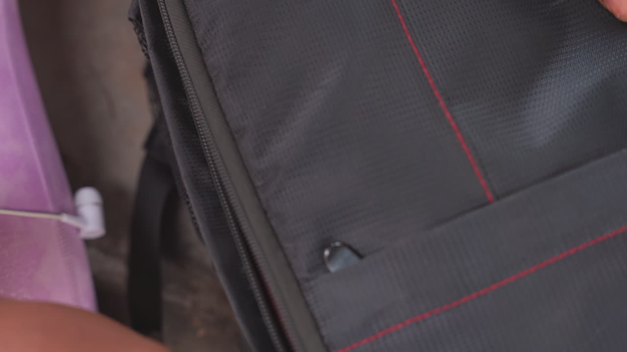 Primer plano cerrado de manos abriendo el bolsillo de una mochila de viaje, un tirón rápido y preciso a lo largo de la línea de costura roja, el deslizador moviéndose sobre nylon duradero, un viajero urbano preparando su equipo, con acceso rápido y embalaje organizado.