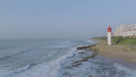 the umhlanga lighthouse on a sunny day with waves crashing against the shore, urban backdrop, aerial view