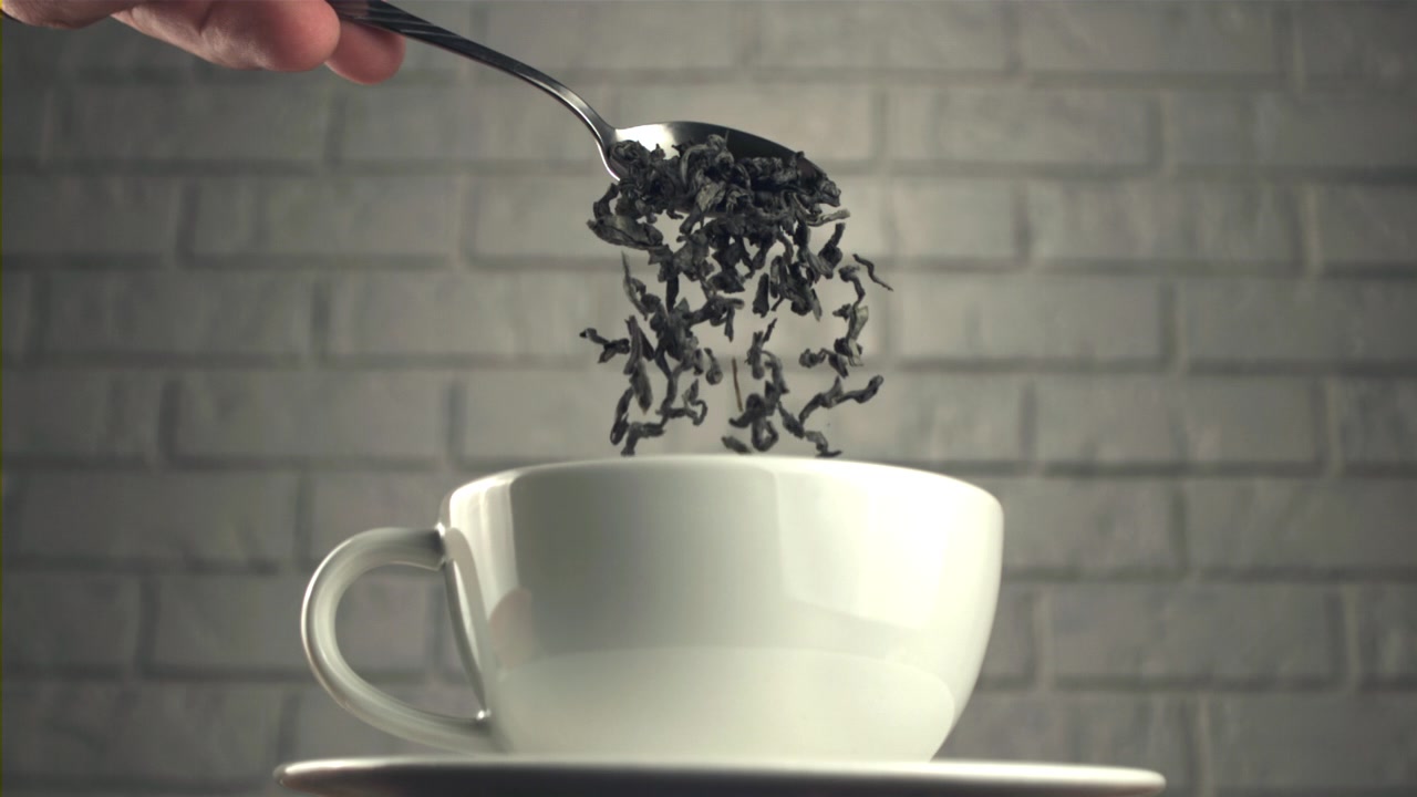 Foglie di tè secche che cadono da un cucchiaio in una tazza bianca al rallentatore