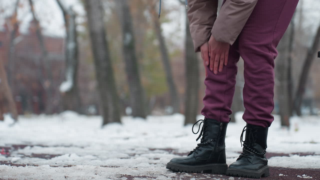 겨울 옷을 입은 젊은이가 무 통증을 느끼고, 구부러지고, 뒷면에 나무가 있는 눈 인 야외 환경에서 스 근처에서 무을 문지르며, 추운 겨울 공원 환경에서 불편함을 나타니다.