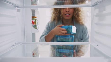 view looking out from inside of refrigerator as woman stacks healthy packed lunches in containers