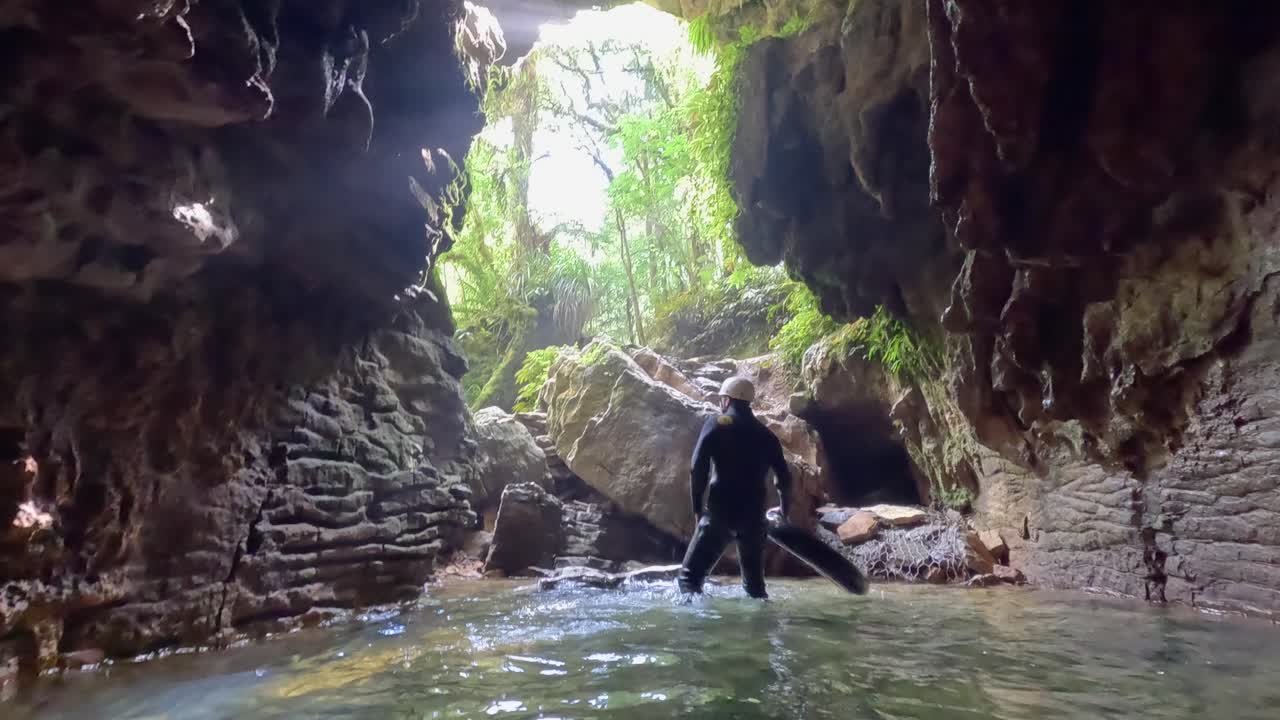 A man emerges from the exit of a rugged cave in Waitomo, New Zealand. Stunning rock formations, stalagmites, and natural scenery highlight the dramatic underground landscape
