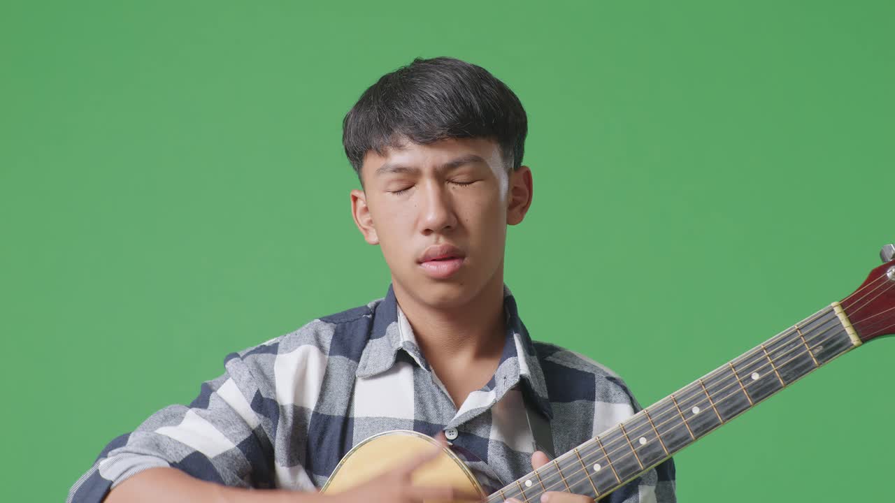 녹색 화면 배경 에 서 있는 동안 두통 을 겪고 있는 기타 를 들고 있는 아시아 청소년 의 근접 사진