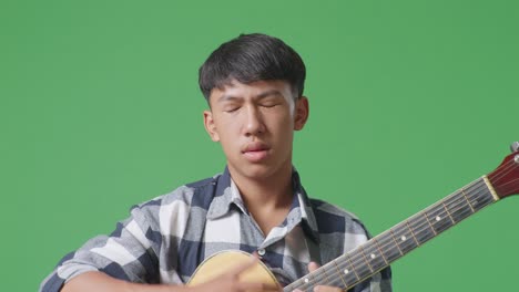 primer plano de un adolescente asiático con guitarra que tiene dolor de cabeza mientras está de pie en el fondo de la pantalla verde