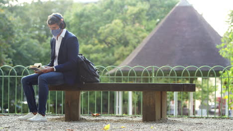 businessman wearing mask reading book and listening on headphones in park with takeaway coffee