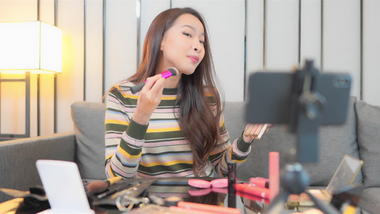 Young woman using cosmetics and applying some make up on her face recommending some beauty products while recording a video on her mobile phone