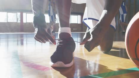 Afroamerikanischer-Männlicher-Basketballspieler,-Der-Schuhe-Mit-Der-Mannschaft-Im-Hintergrund-Bindet