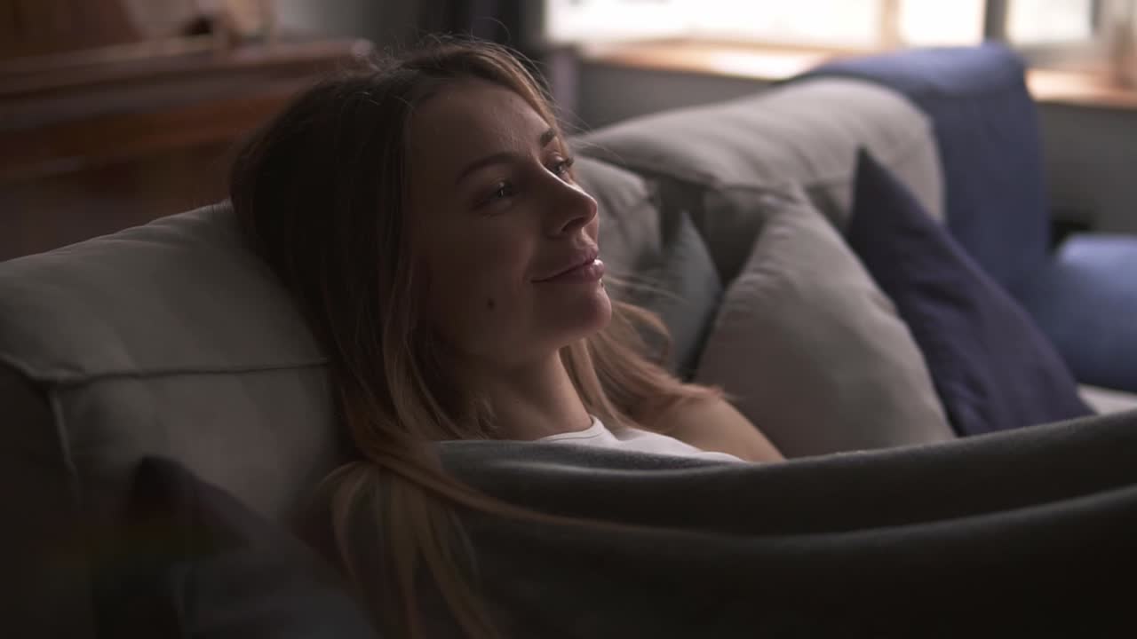 소파에 앉아 있는 미소 짓는 아름다운 여성은 좋은 무언가를 꿈꾸고, 플레이드로 싸여 있습니다.