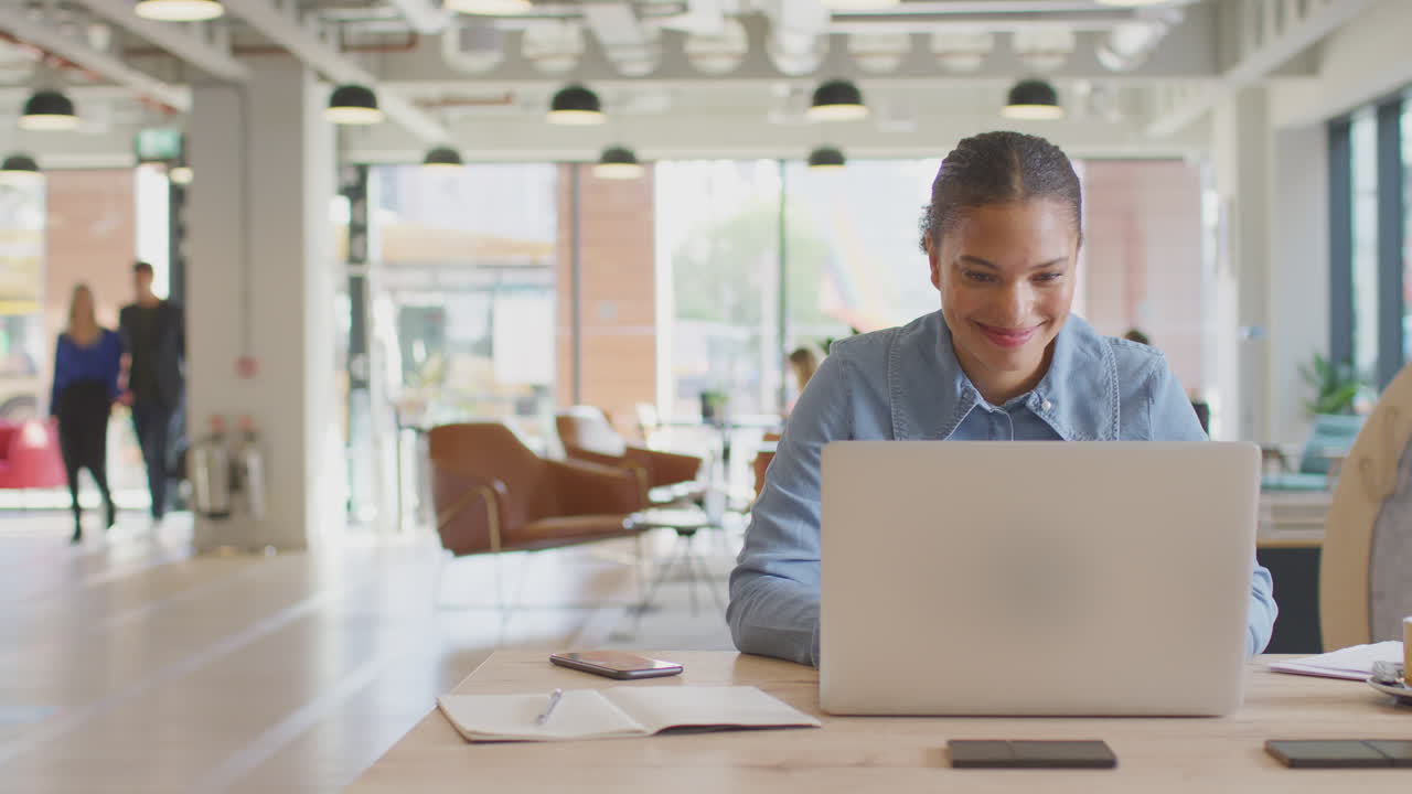 Young Businesswoman Working On Laptop At Desk In Modern Office With Colleagues In Background