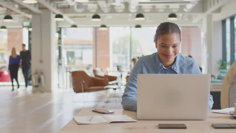 young businesswoman working on laptop at desk in modern office with colleagues in background