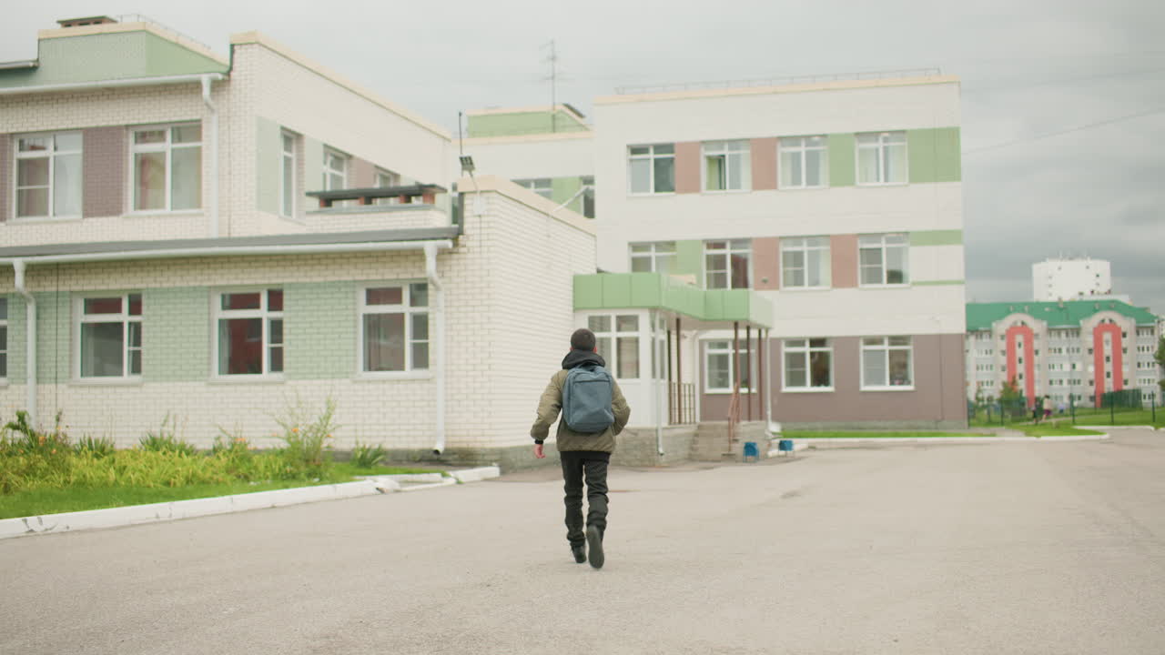 배낭을 메고 학교 건물 쪽으로 달려가다 옆으로 시선을 돌리는 남자아이의 뒷모습이에요. 흐린 하늘 아래 새가 길을 가로질러 날아가고, 배경에는 다채로운 주거 건축물이 있는 도시 동네의 모습이 담겨 있어요.