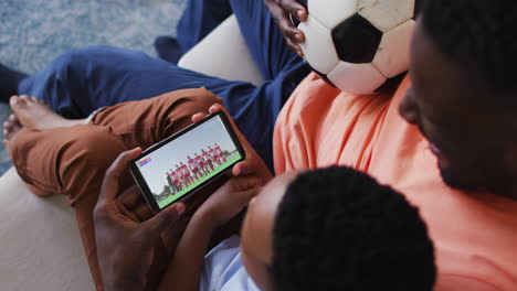 composite of father and son at home watching sports event on smartphone